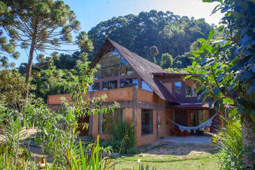 A modern, rustic house with large windows and a slanted roof, surrounded by lush greenery and tall trees. A hammock hangs on the covered porch.