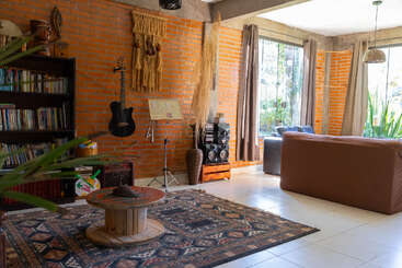 A cozy living room with brick walls, large windows, brown sofa, bookcase, guitar, stereo, decorative rug, and natural light. Plants and earthy decor create warmth.