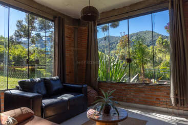Sunlit room with brick walls, large glass windows, leather sofa, and a small wooden table with a plant, overlooking lush greenery and scenic mountains outside.
