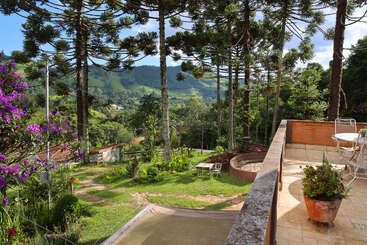 A scenic terrace overlooks a lush, green landscape with tall trees, distant mountains, flowering bushes, and outdoor seating, creating a peaceful and inviting atmosphere.