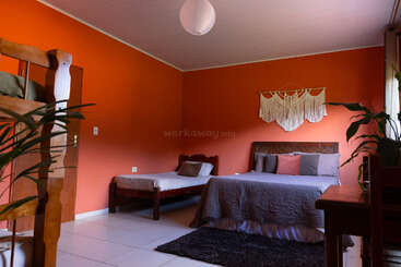 The room features orange walls, a double bed, single bed, bunk bed, indoor plants, a Macramé wall hanging, desk, and white tiled floor. Cozy atmosphere.
