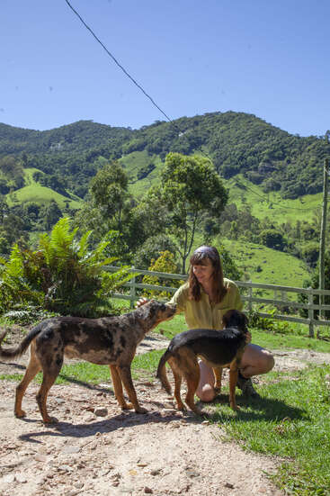 A woman kneels on a dirt path, petting two dogs with a white fence and rolling hills in the background on a sunny day.