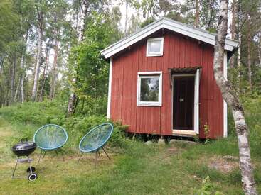 Eine kleine rote Holzhütte steht in einem üppigen grünen Wald. Auf dem grasbewachsenen Rasen stehen zwei blaue Stühle und ein schwarzer Grill, die zum Entspannen einladen.