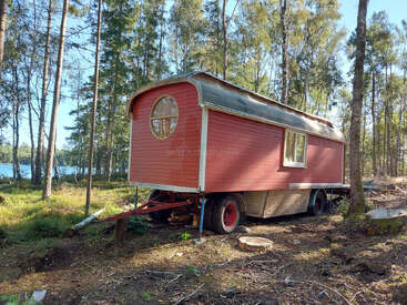 Ein rotes mobiles Holzhäuschen mit einem runden Fenster steht auf einer Waldlichtung in der Nähe eines Sees, umgeben von hohen Bäumen und Sonnenlicht. Friedliche Umgebung.