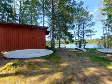 Ein roter Holzschuppen steht zwischen hohen Kiefern am Ufer eines ruhigen Sees. Mehrere Kajaks oder Kanus liegen auf dem Boden und bilden eine friedliche, rustikale Szene.