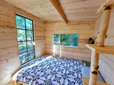 Ein gemütliches Schlafzimmer in einer Holzhütte hat große Fenster mit Blick auf den Wald, eine gemusterte Bettdecke, eine Einrichtung aus Naturholz und ein einzigartiges Regal aus Birkenholz.