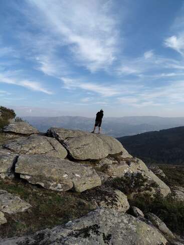 Una persona está de pie sobre unas grandes rocas, contemplando un vasto paisaje montañoso. El cielo es azul con nubes difusas, creando una atmósfera serena y contemplativa.