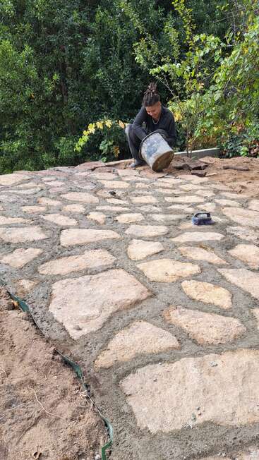 Una persona está colocando grandes baldosas de piedra al aire libre, rellenando huecos con cemento. Rodeados de vegetación, trabajan con cuidado, sujetando un cubo sobre una superficie recién pavimentada.