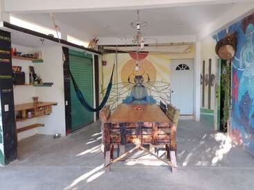 A sunlit room with a wooden table and chairs, hammock, colorful wall murals, hanging light fixture, open shelves, and a white door create an artistic atmosphere.