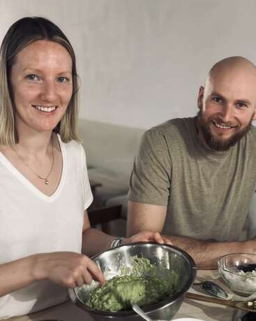 Uma mulher e um homem sorridentes sentados em uma mesa. A mulher está mexendo guacamole verde em uma tigela de metal, sugerindo que eles estão preparando comida juntos em uma atmosfera amigável.