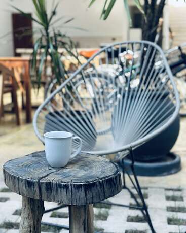 Uma caneca branca fica em uma mesa de madeira rústica. Atrás dela, uma moderna cadeira de arame e plantas verdes criam um ambiente externo aconchegante e descontraído com luz natural.
