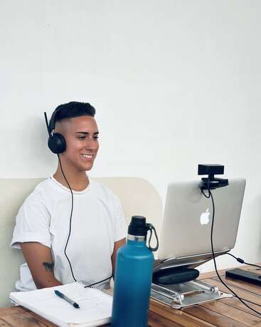 Uma pessoa usando fones de ouvido sorri durante uma chamada de vídeo, sentada em uma mesa com um laptop, garrafa de água, caderno, caneta e webcam, em um fundo branco liso.