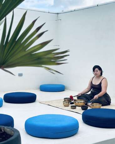 Uma mulher senta-se de pernas cruzadas em um tapete em um espaço externo minimalista, tocando tigelas de canto. Almofadas azuis e uma grande planta criam uma atmosfera tranquila.