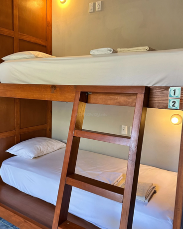 Esta imagem mostra um beliche de madeira com roupa de cama branca, camas numeradas, uma escada de madeira, luzes de parede, tomadas elétricas e toalhas bem dobradas em cada colchão.