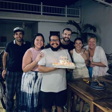 Um grupo de seis amigos comemora alegremente um aniversário dentro de casa. O aniversariante segura um bolo com velas de "Feliz Aniversário". Todos sorriem, irradiando calor e felicidade juntos.