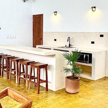 A imagem mostra uma cozinha moderna com paredes e bancadas brancas, com uma pia, um micro-ondas e banquetas de bar, em um piso amarelo e um vaso de plantas.