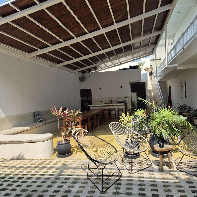 A imagem mostra uma sala espaçosa com teto alto, com uma escada, plantas e cadeiras, com um homem descendo as escadas ao fundo.