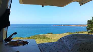 El acogedor interior de la furgoneta ofrece una impresionante vista del mar, el océano azul y el cielo se encuentran con los verdes acantilados, y en su interior se ven un lavabo, una cama y aperitivos.