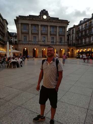 La imagen muestra a un hombre de pie delante de un gran edificio con un reloj, rodeado de gente y edificios al fondo, en un cielo nublado.