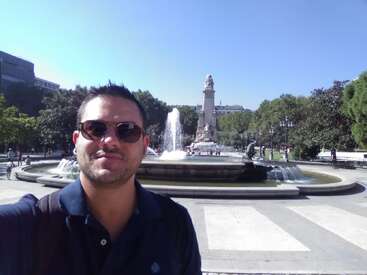 La imagen muestra a un hombre delante de una fuente, con un alto monumento al fondo, rodeado de árboles y un cielo azul despejado.