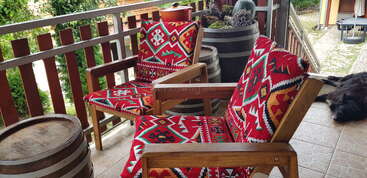 Deux chaises en bois avec des coussins aux motifs vibrants et colorés sont assises sur un balcon carrelé avec des tonneaux en guise de tables. Un chien noir se détend près de la balustrade du balcon.
