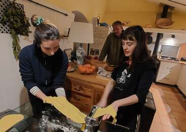 Three women and a man make homemade pasta in a cozy kitchen. They shape ravioli, roll dough, and enjoy cooking together, creating a warm, communal atmosphere.