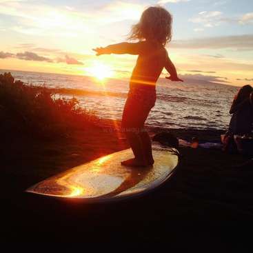 La imagen muestra a una joven surfeando en una tabla al atardecer, con los rayos del sol brillando a través de su cuerpo. La silueta de la chica se recorta contra el cielo.