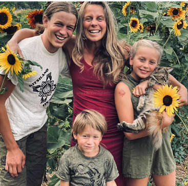 Una mujer sonriente con tres hijos posa en un campo de girasoles. La niña sostiene un gato y un girasol, creando un momento familiar alegre, cálido y lleno de naturaleza.