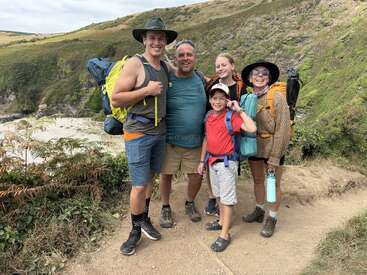 Cinco personas sonrientes posan con sus mochilas en una ruta de senderismo con vistas panorámicas. Están rodeadas de colinas verdes y rocosas, y todos parecen felices y listos para la aventura.