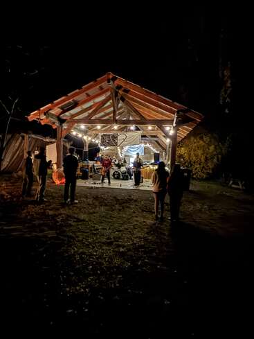 Bei einem nächtlichen Open-Air-Konzert spielt eine kleine Band unter einem Holzpavillon, der von Lichterketten warm beleuchtet wird, während ein paar Leute dastehen und zuschauen.