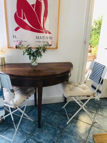 Um cantinho aconchegante com uma mesa de madeira, duas cadeiras brancas almofadadas, piso de azulejos azuis, um vaso com flores, uma gravura de Matisse e a luz do sol entrando pela janela.