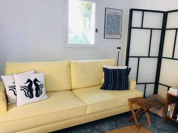 Uma aconchegante sala de estar com um sofá amarelo claro, almofadas com tema náutico, uma almofada azul, um biombo, uma janela, quadros, uma mesinha e uma estante.