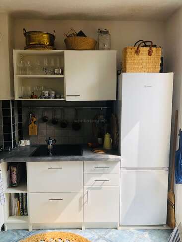 Essa cozinha pequena e aconchegante tem armários brancos, uma pia preta, uma geladeira, prateleiras abertas com copos, cestas, uma panela de latão, livros, utensílios e luz natural.