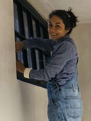 Uma mulher sorridente, vestindo um macacão jeans e uma camisa xadrez, trabalha na moldura preta de uma janela dentro de um cômodo de cores claras, curtindo seu projeto de bricolagem.
