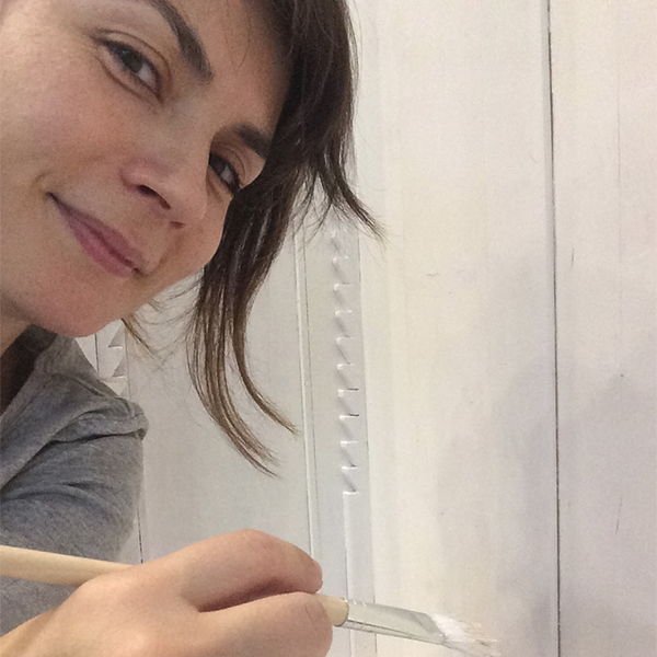 The image depicts a woman with dark hair, wearing a gray shirt, holding a paintbrush and painting a white wall, likely in a DIY or creative context.