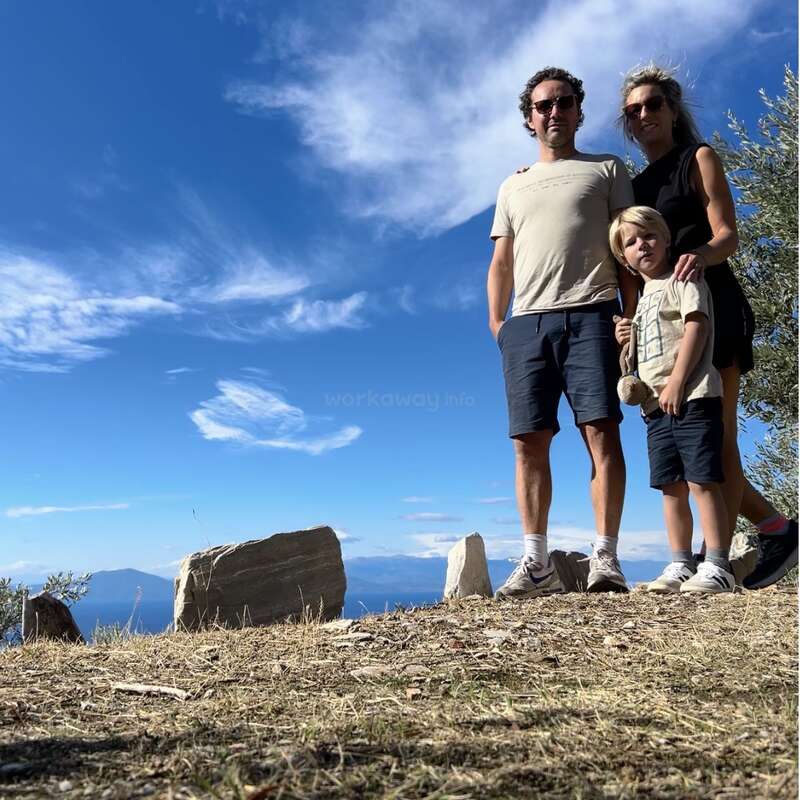 Uma família de três pessoas está ao ar livre, em um terreno rochoso, sob um céu azul brilhante com nuvens espalhadas, aproveitando um dia ensolarado juntos em um cenário natural pitoresco.