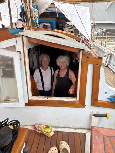 An older couple stands smiling inside the cozy cabin of a wooden sailboat. Shoes rest outside the entrance. Nautical ropes, gear, and personal items surround the scene.