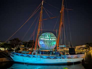 A sailboat docked at night displays a glowing projection reading "Virtual Saga" on its sail. The scene features colorful lights and a festive atmosphere.