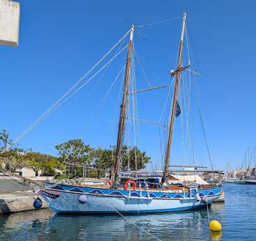 A colorful sailboat with two tall masts is docked at a marina on a bright, sunny day. The water is calm, sky clear blue.