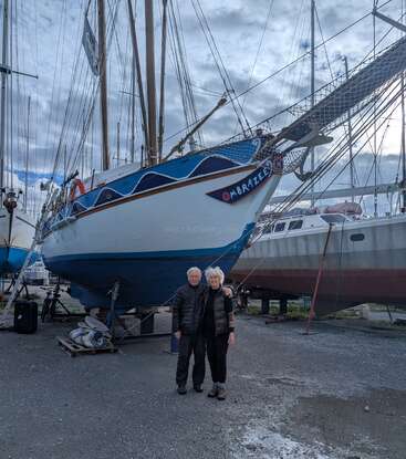 An older couple stands arm-in-arm, smiling, in front of a large blue-and-white sailboat named "Ombrazee," which is dry-docked among other boats. Overcast sky.