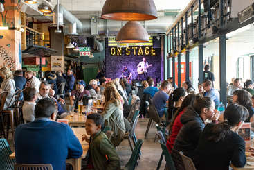 Das Bild zeigt eine lebhafte Szene in einem Restaurant oder einer Bar mit einer Bühne und einem Publikum, in dem eine Gruppe von Menschen versammelt ist, die wahrscheinlich eine Aufführung oder eine Veranstaltung genießen.