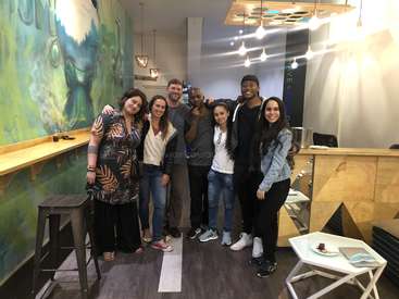 Das Bild zeigt eine Gruppe von sieben Personen, die für ein Foto in einem modernen Café posieren, umgeben von einem Wandgemälde, Barhockern und einem Tresen mit Hängelampen.