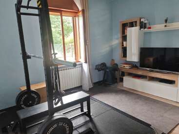 Das Bild zeigt einen Raum mit hellblauen Wänden, in dem ein Hantelständer, eine Bank, ein Fernseher, Regale, Trainingsgeräte und ein offenes Fenster mit Vorhängen zu sehen sind.