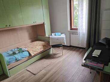 Dieses Bild zeigt ein aufgeräumtes Schlafzimmer mit einem Einzelbett, grünen Schränken, einem Fenster mit transparenten Vorhängen, einem Schreibtisch mit Computer, einem Stuhl und einem Holzboden.