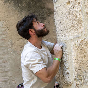 Um homem de barba e cabelo curto, vestindo uma camiseta branca e uma pulseira verde, prepara-se para escalar uma parede de pedra irregular, com um ar determinado.