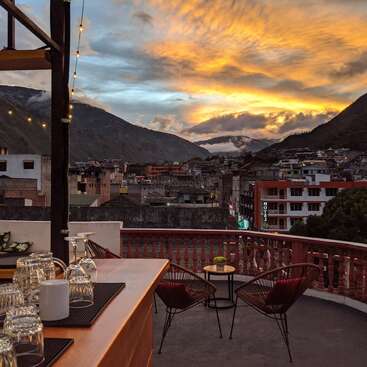 Das Bild zeigt eine Rooftop-Bar mit einem atemberaubenden Blick auf eine Bergstadt bei Sonnenuntergang, mit einer Bar, Stühlen und Tischen und einem leuchtenden Himmel mit Wolken.
