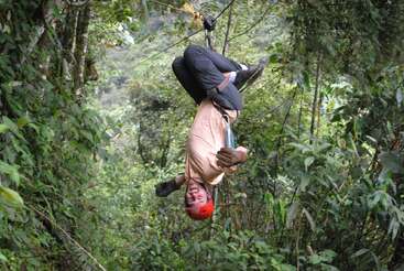 Una persona, con un casco y guantes rojos, cuelga boca abajo de una tirolina en medio de un frondoso bosque, con aire aventurero y emocionado, disfrutando de la actividad al aire libre.