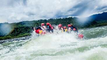 Un grupo de personas con cascos rojos y chalecos salvavidas practican rafting en aguas bravas por los rápidos de un río agitado, rodeados de exuberantes colinas verdes y un cielo nublado de aspecto espectacular.