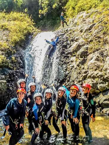 Un grupo de personas sonrientes, con cascos y trajes de neopreno, posa junto a una pequeña cascada, señalando a alguien que desciende en rapel por las rocas, disfrutando de una excursión llena de aventura.