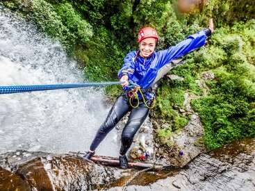 Una mujer vestida con ropa de aventura y casco sonríe mientras desciende en rapel por una cascada, rodeada de un frondoso bosque, sujetando una cuerda azul y disfrutando de la emoción.